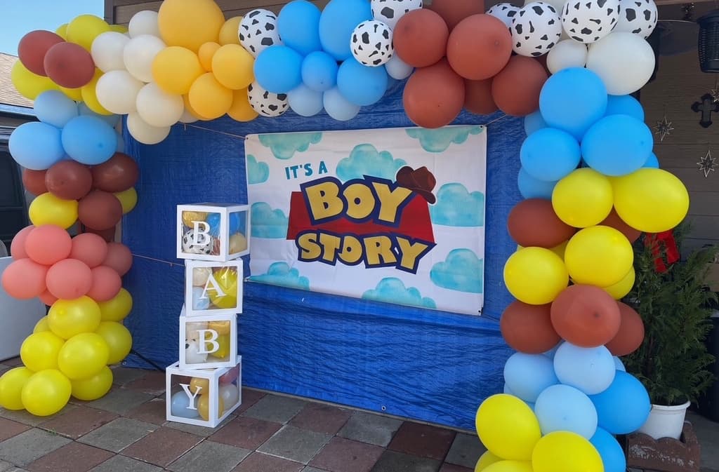 Arco de globos amarillos, azules, color óxido y con estampado de vaca alrededor de un letrero de "It's a Boy Story", con cubos de letras que dicen BABY