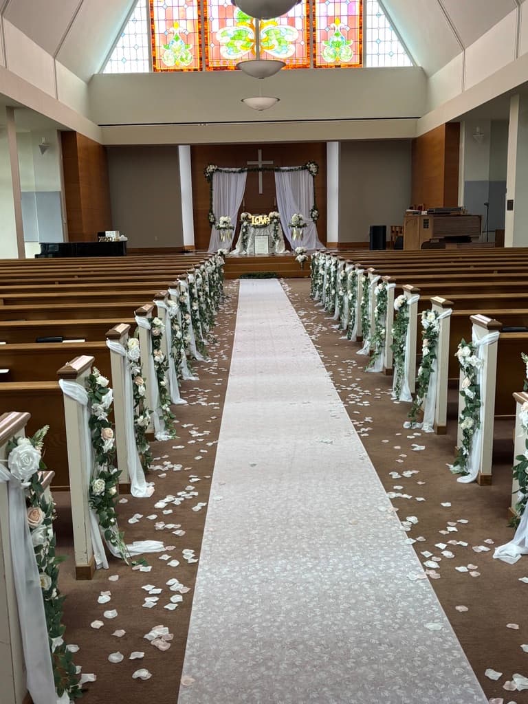 Pasillo de iglesia preparado para una boda, con un camino blanco, pétalos esparcidos y guirnaldas de flores en cada banca