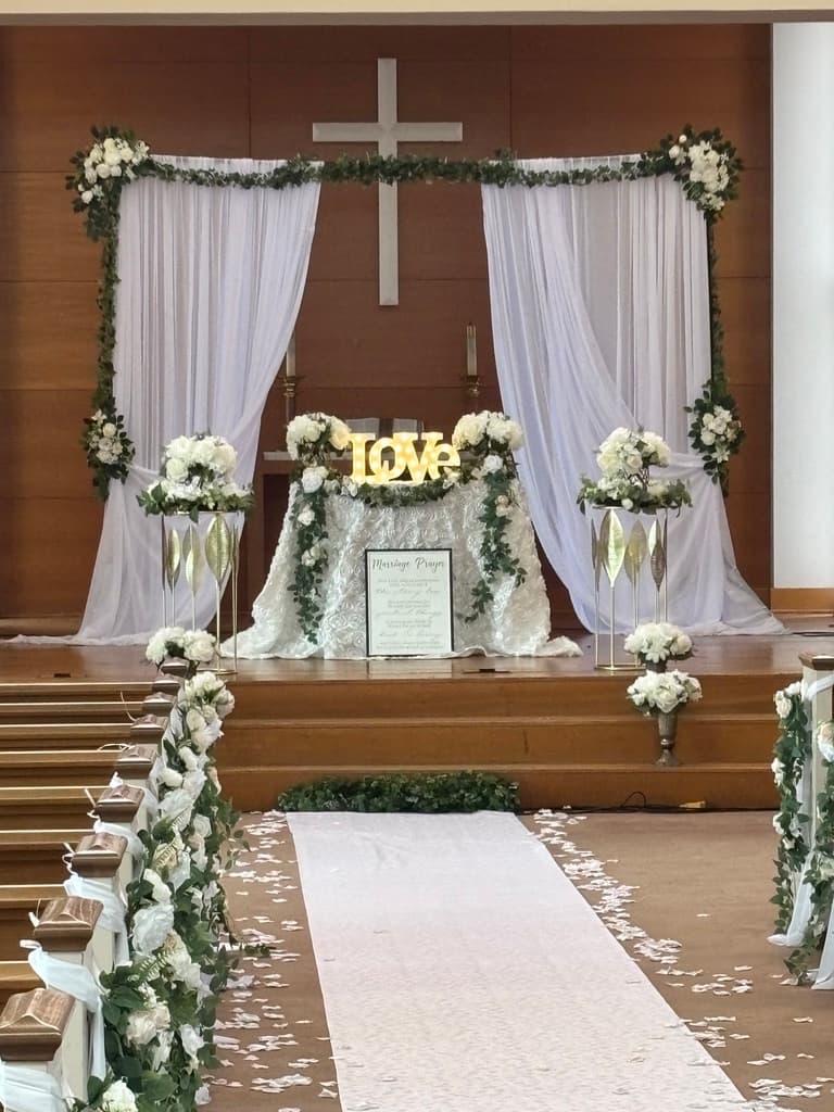 Altar de iglesia decorado para una boda: cortinas blancas y follaje alrededor de un letrero iluminado de LOVE, bases con flores blancas y un camino de pasillo blanco