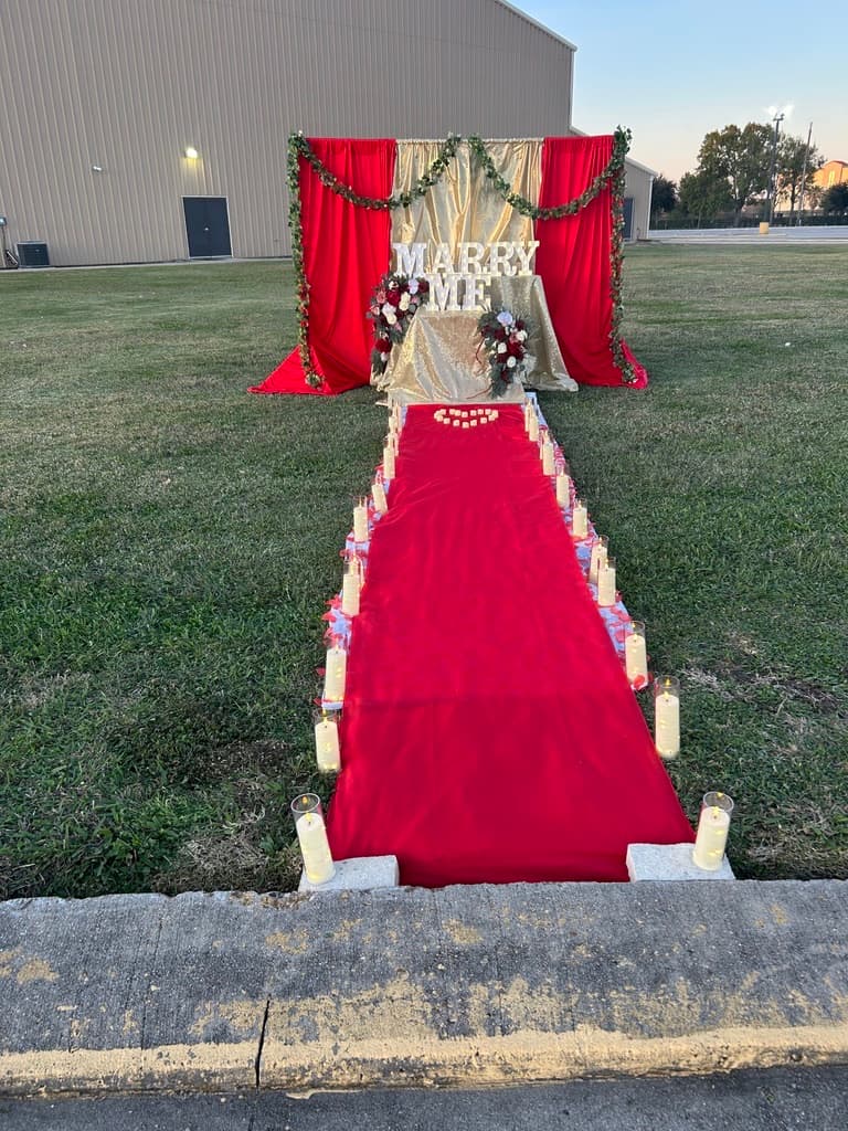 Decoración al aire libre para una propuesta de matrimonio: una alfombra roja bordeada de velas que lleva a un letrero iluminado de MARRY ME bajo cortinas rojas y doradas y una guirnalda de follaje