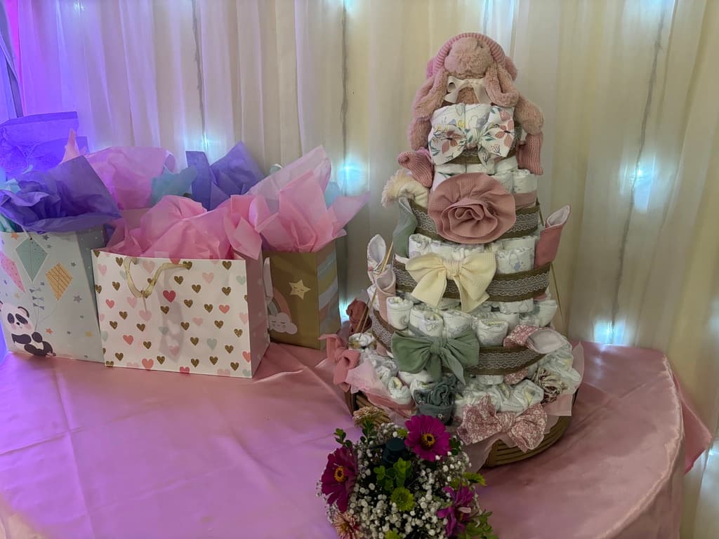 Pastel de pañales rosa con un peluche de conejito y moños de tela, junto a bolsas de regalo en una mesa rosa