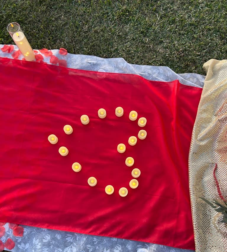 Corazón de velitas sobre un camino rojo, con pétalos de rosa y una vela de pilar sobre el pasto