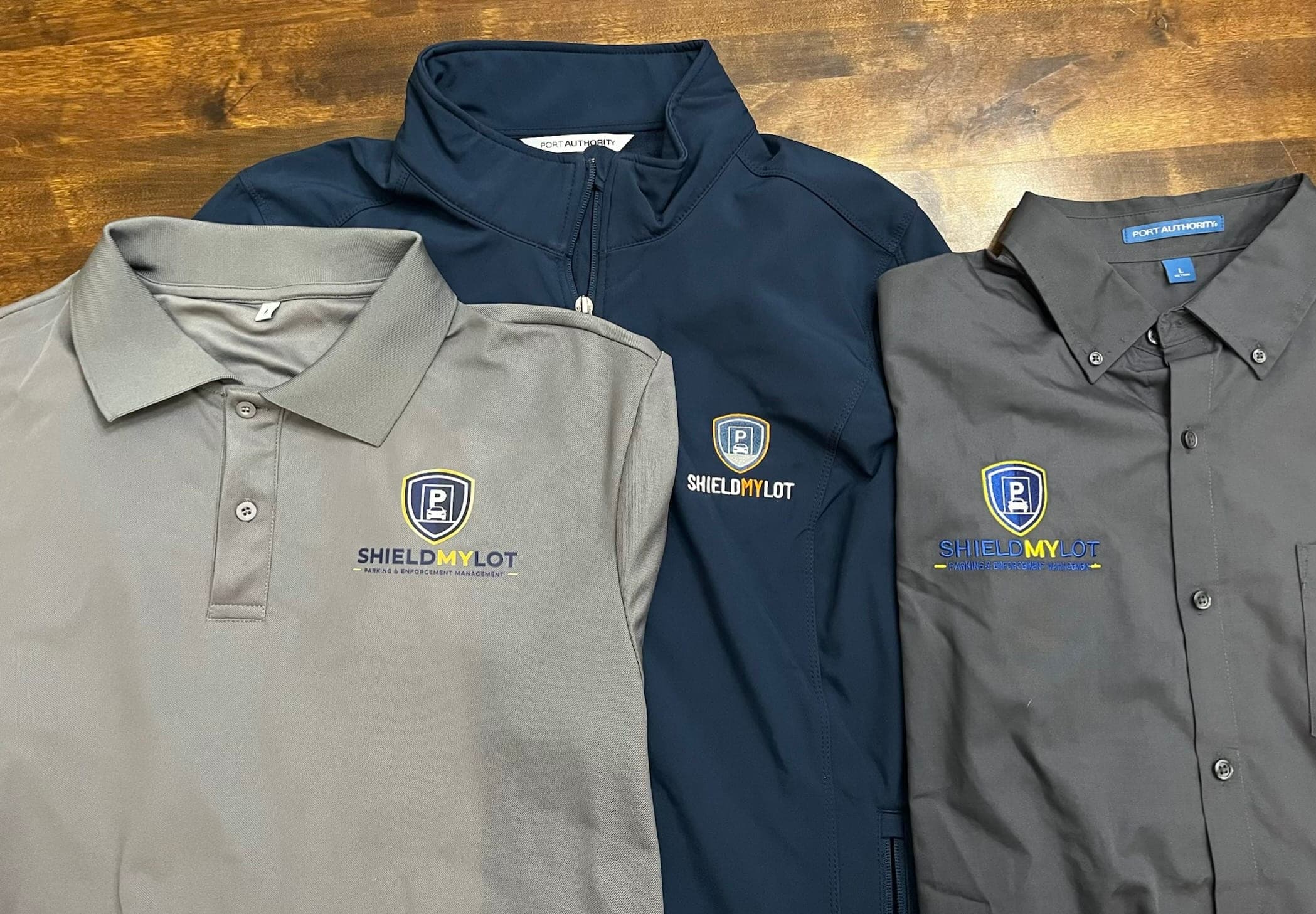 Ropa personalizada que S&A hizo para el equipo de ShieldMyLot: una playera polo gris con el logo de ShieldMyLot impreso en el pecho, una chamarra azul marino y una camisa de vestir gris oscuro con el logo bordado