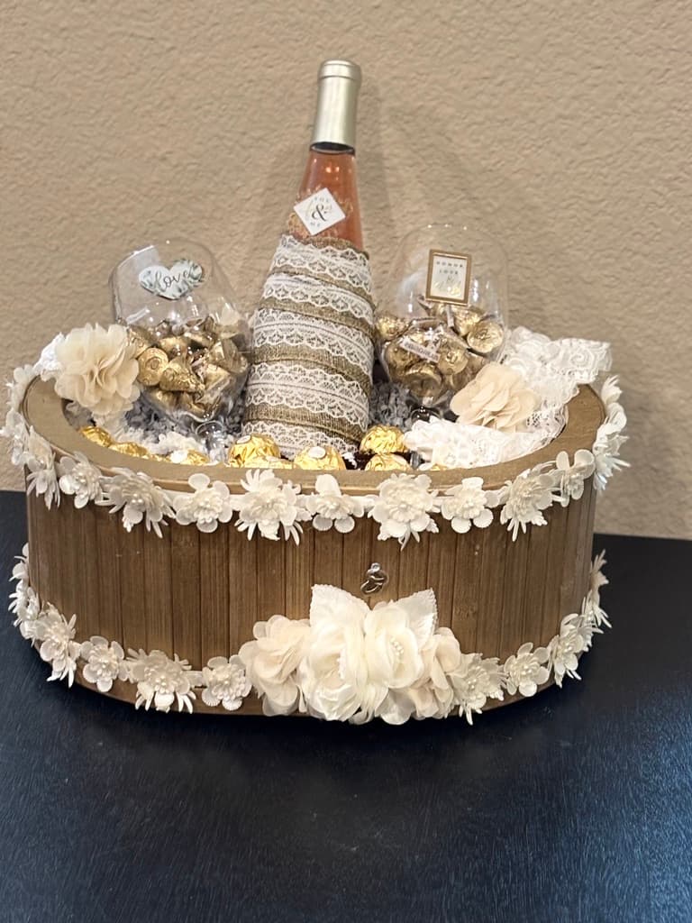 Canasta de regalo de boda con una botella de vino rosado envuelta en encaje, dos copas llenas de chocolates y adornos de flores color marfil