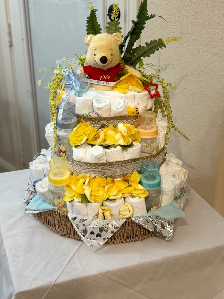 Pastel de pañales de tres pisos con un peluche de Winnie the Pooh arriba, adornado con flores amarillas, yute y biberones